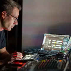 Mężczyzna w okularach pracuje przy konsoli dźwiękowej i laptopie, tworząc muzykę w swoim domowym clubhouse.