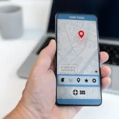 Dłoń trzyma smartfon z aplikacją do śledzenia telefonu. Na ekranie widoczna jest mapa z pinezką lokalizacji i przyciskiem SOS.
