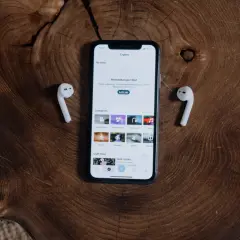 Jak połączyć AirPods z iPhone? Na drewnianym blacie leży iPhone z otwartą aplikacją i dwa białe słuchawki AirPods.