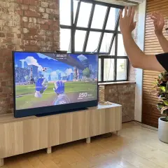 Mężczyzna gra w grę wideo na telewizorze, na którym widać wirtualne rękawice bramkarskie. Obraz na ekranie przypomina niebo i szkło.