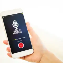 Dłoń trzyma smartfon z włączonym dyktafonem w telefonie. Na ekranie widnieje komunikat "VOICE MESSAGE" i przycisk "Stop".