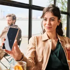 Kobieta trzyma telefon, słuchając wiadomości na poczcie głosowej. W tle mężczyzna i widok na port.