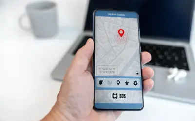 Dłoń trzyma smartfon z aplikacją do śledzenia telefonu. Na ekranie widoczna jest mapa z pinezką lokalizacji i przyciskiem SOS.