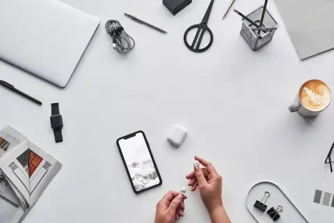 Ręce trzymające słuchawki AirPods obok telefonu z Androidem, laptopa i kawy. Czy AirPods działają z Androidem? Tak!