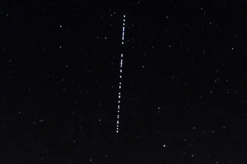 Nocne niebo z gwiazdami i pionowym sznurkiem jasnych punktów, przypominających przeloty Starlink.