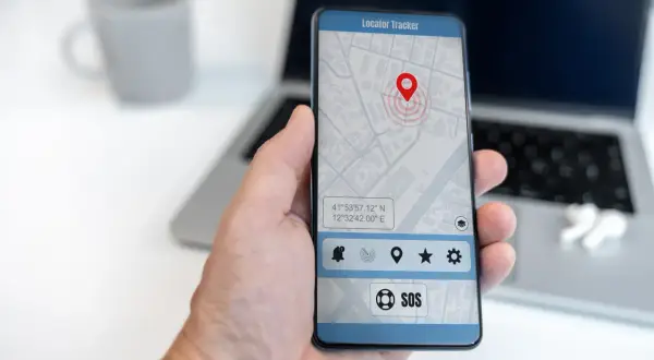 Dłoń trzyma smartfon z aplikacją do śledzenia telefonu. Na ekranie widoczna jest mapa z pinezką lokalizacji i przyciskiem SOS.