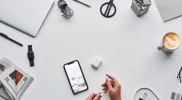 Ręce trzymające słuchawki AirPods obok telefonu z Androidem, laptopa i kawy. Czy AirPods działają z Androidem? Tak!