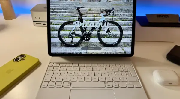 Na ekranie iPada z systemem ipados 26 widoczny jest rower szosowy z napisem "witamy". Obok leży żółty iPhone i biała klawiatura.