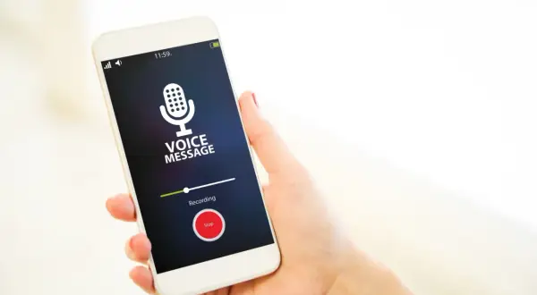 Dłoń trzyma smartfon z włączonym dyktafonem w telefonie. Na ekranie widnieje komunikat "VOICE MESSAGE" i przycisk "Stop".