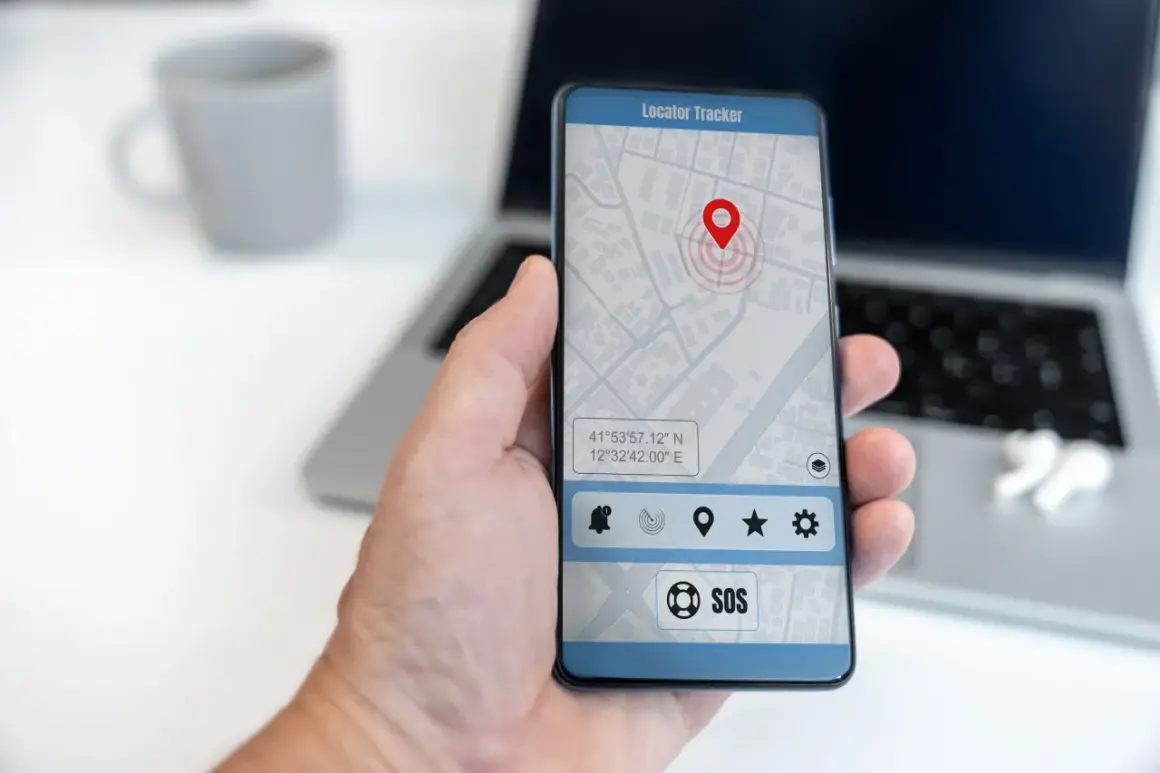 Dłoń trzyma smartfon z aplikacją do śledzenia telefonu. Na ekranie widoczna jest mapa z pinezką lokalizacji i przyciskiem SOS.