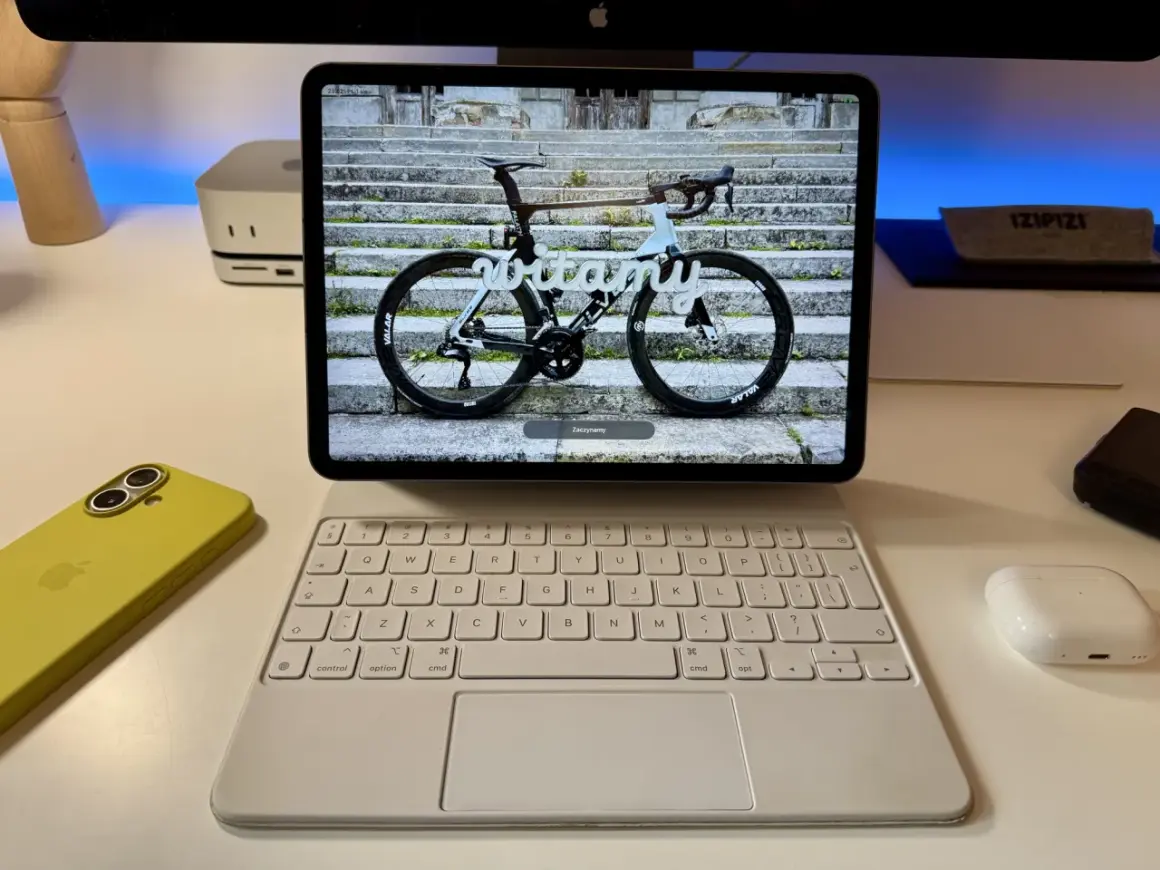 Na ekranie iPada z systemem ipados 26 widoczny jest rower szosowy z napisem "witamy". Obok leży żółty iPhone i biała klawiatura.