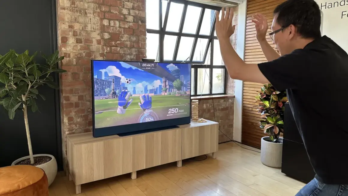 Mężczyzna gra w grę wideo na telewizorze, na którym widać wirtualne rękawice bramkarskie. Obraz na ekranie przypomina niebo i szkło.