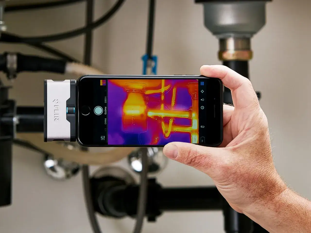 Dłoń trzyma smartfon z podłączonym modułem termowizyjnym. Na ekranie telefonu widoczny obraz termowizyjny instalacji hydraulicznej, pokazujący różnice temperatur.