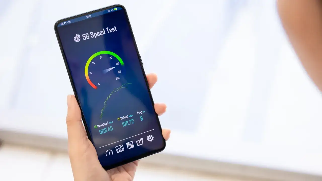 Dłoń trzyma smartfon z aplikacją do testowania prędkości 5G. Widoczne są wyniki pobierania, wysyłania i ping, świadczące o szybkich danych komórkowych.