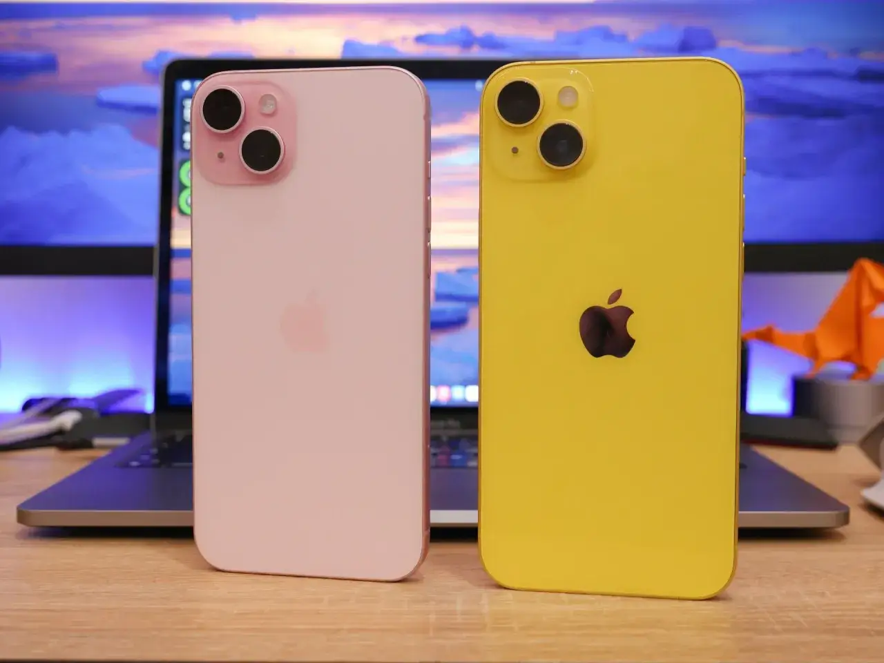 Różowy i żółty iPhone, który wybrać? iPhone 14 czy 15? Dwa telefony na biurku, obok laptopa.