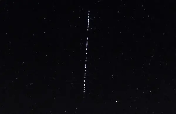 Nocne niebo z gwiazdami i pionowym sznurkiem jasnych punktów, przypominających przeloty Starlink.