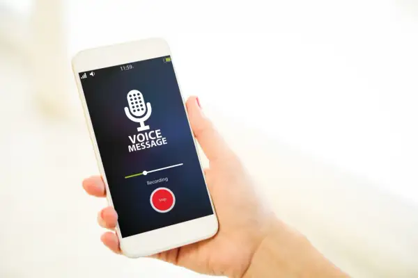 Dłoń trzyma smartfon z włączonym dyktafonem w telefonie. Na ekranie widnieje komunikat "VOICE MESSAGE" i przycisk "Stop".